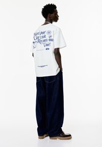 Hvid grafisk t-shirt med blå tekst og design på ryggen, parret med løst siddende mørke denim jeans og brune platform sko.