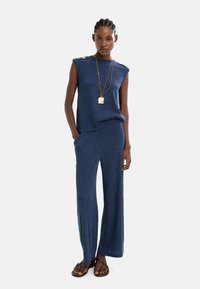 Granatowy dzianinowy kombinezon z bezrękawnikami, bocznymi kieszeniami i szerokimi nogawkami. Złote guziki akcentujące na ramionach, zestawione z płaskimi sandałami.