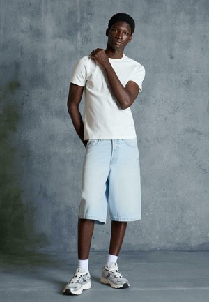 Fiatal férfi, fehér pólóban, világoskék farmer rövidnadrágban, fehér zokniban és vastag talpú sportcipőben, szürke texturált fal előtt állva.