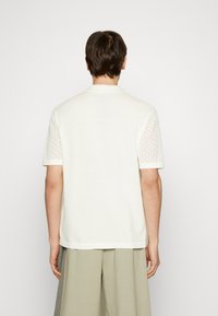 Polo de manga corta crema con patrón de malla texturizada en los hombros, cuello de punto plano y corte ajustado, combinado con pantalones sueltos gris claro.