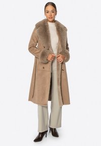 Abrigo largo de piel de oveja color beige con cuello y puños de piel, con un corte entallado y botones delanteros, combinado con un suéter de cuello alto color crema y pantalones beige.