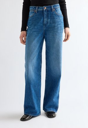 Persona che indossa jeans blu a vita alta e gamba larga, top nero a maniche lunghe e scarpe nere con punta quadrata, in piedi su sfondo bianco.