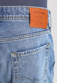 Vaaleansiniset farkut, joissa on nahkainen merkki, jossa lukee "JACK & JONES", näkyvää ommeltua saumaa ja takataskun yksityiskohta.