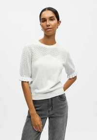 Pull en tricot blanc avec des manches en maille à motif losange, des poignets volants et une coupe droite. Associé à un jean gris taille haute.