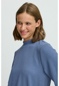 Modra rebrasta majica s kapuco in visokim ovratnikom, textured material in dolgimi rokavi. Šivi so vidni na ramenih. Mehka, sproščena prileganje.