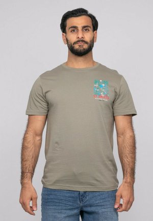 Homme aux cheveux foncés et barbe, portant un t-shirt vert olive avec un petit motif tropical bleu et rouge, associé à un jean bleu, debout devant un fond gris.