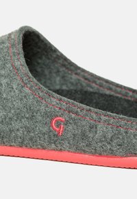 Grå følesko med et rundet slip-on design, kontrasterende rød syning og en lys rød gummisål. Har et lille broderet "G."