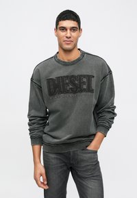 Sweatshirt cinza com decote redondo e textura desgastada, apresentando um atrevido logo preto "DIESEL" e punhos e barra canelados. Combinado com jeans escuros.