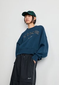 Kék kapucnis pulóver "NIKE" felirattal, vastag betűtípussal, fekete sportnadrággal, fehér díszítéssel és logóval párosítva. Fekete és zöld kockás sapka.