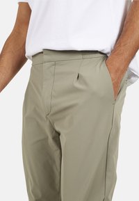 Jasnokhaki spodnie o gładkiej fakturze, z bocznymi kieszeniami i płaskim fasonem. Noszone z prostą białą koszulką.