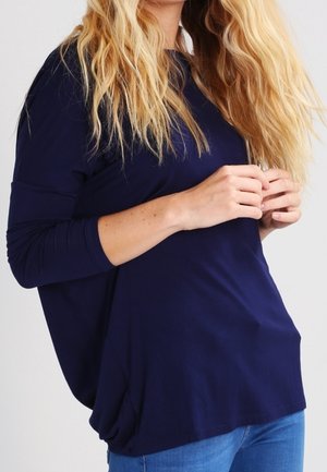 Kvinde med langt blondt hår iført en løs mørkeblå langærmet skjorte og blå jeans, der holder sin skjorte nær brystet.