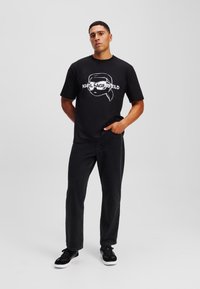 Camiseta negra con un diseño gráfico blanco que presenta un personaje de dibujos animados y el texto "KARL LAGERFELD." Combinada con pantalones vaqueros negros y zapatillas.