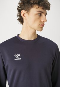 Maglietta a maniche lunghe blu navy realizzata in tessuto morbido, con scollatura a girocollo e logo bianco stampato sul petto. Tessuto liscio con design minimal.