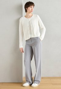 Weißer, gestrickter Cardigan mit strukturiertem Muster, kombiniert mit hellgrauen, weiten Hosen und weißen Sneakers, vor einem neutralen Hintergrund.