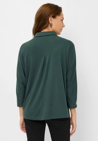 Donkergroene, lichte blouse met een kraag en driekwartmouwen. Beschikt over een rugplooitje en een gladde textuur. Eenvoudig, ongecompliceerd ontwerp.