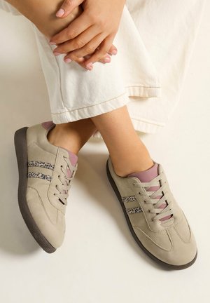 Personne portant des baskets beige avec des accents roses et des rayures imprimé léopard, assise les mains jointes sur les jambes croisées.