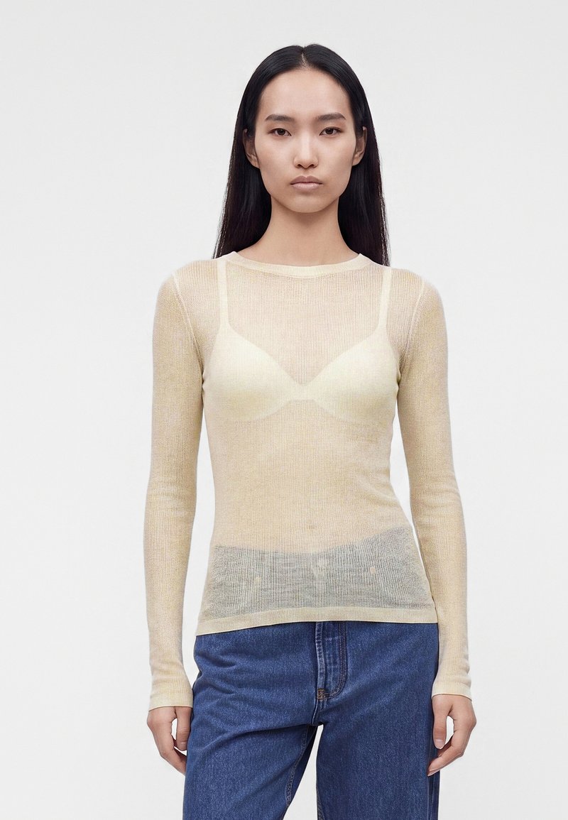 Jeune femme aux longs cheveux foncés porte un top beige transparent à manches longues par-dessus un soutien-gorge crème, associé à un jean bleu taille haute.