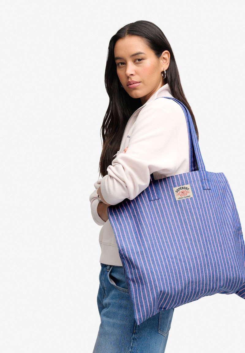 Woman with long dark hair carrying large blue tote bag with pink stripes and logo, wearing light jacket and blue jeans, standing sideways.