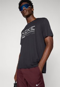 Fekete Nike póló szürke logóval és csíkos dizájnnal, bordó rövidnadrággal párosítva. Reflektív kék napszemüveg. Sima textúra, laza szabás.