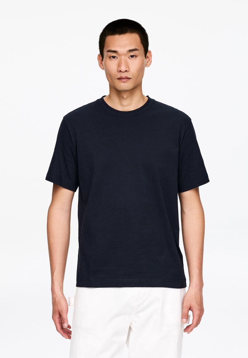 Jeune homme aux cheveux courts noirs portant un t-shirt noir uni à manches courtes et un pantalon blanc, debout devant un fond blanc.