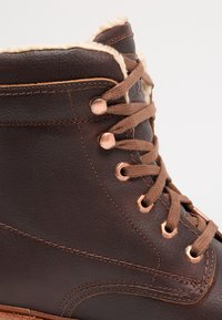 Bota de tornozelo em pele castanha com colarinho em pêlo sintético, ilhóses de cobre e atacadores castanhos. Superfície texturizada com detalhes de costura em contraste.