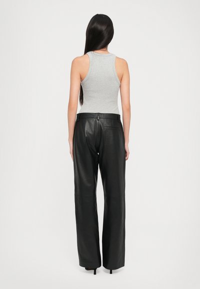 Woman with long dark hair wearing a gray sleeveless top and black wide-leg leather pants, standing facing away on a plain white background.