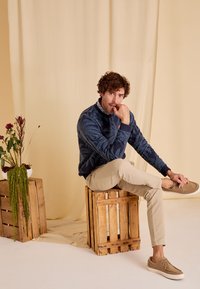Sweatshirt marine avec motif de feuilles de palmier, pantalon beige fuselé et baskets beiges ; modèle assis sur des caisses en bois avec des plantes en arrière-plan.
