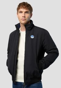 Schwarze Zip-Up-Jacke mit hohem Kragen, gestrickten Bündchen und einem Logo-Patch auf der Vorderseite. Über einem weißen Hemd getragen, zeichnet sie sich durch eine glatte Textur und eine strukturierte Passform aus.