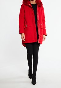 Parka rouge avec capuche bordée de fourrure, dotée d'une fermeture éclair à l'avant, de deux poches latérales et d'un ourlet arrière légèrement plus long. Portée avec des leggings noirs.