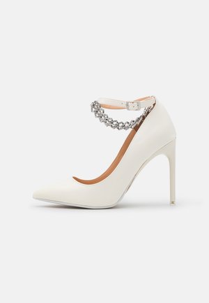Zapato de tacón alto blanco puntiagudo con una cadena plateada y hebilla en la correa del tobillo sobre un fondo claro y sencillo.