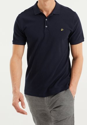 Homme portant un polo à manches courtes bleu marine avec un petit logo banane jaune sur la poitrine gauche et un pantalon gris rayé. Une main dans la poche.