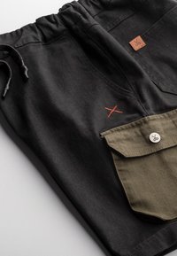 Pantalon noir avec une taille à cordon, présentant une poche vert olive et un détail brodé rouge. Comprend une petite étiquette en cuir de marque.