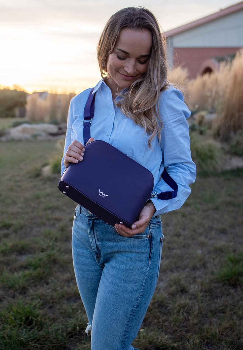 Lila Umhängetasche mit glatter Textur, runder Form und Logo auf der Vorderseite. Verfügt über einen verstellbaren Trageriemen und silberne Beschläge.