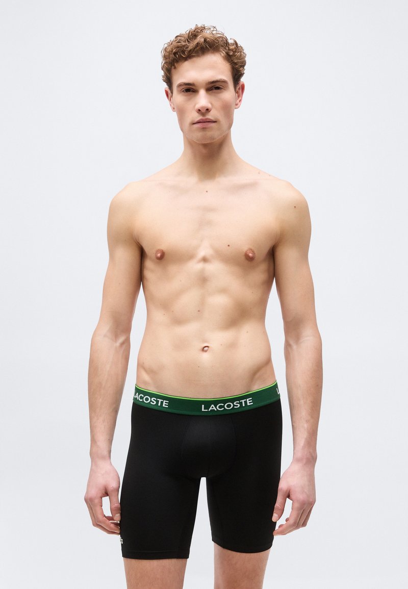 Νεαρός άνδρας με σγουρά μαλλιά στέκεται προς τα εμπρός φορώντας μαύρα εσώρουχα Lacoste με πράσινη μέση σε απλό φωτεινό φόντο.