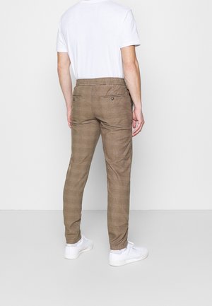 Man in bruin geruite broek, witte t-shirt en witte sneakers staat met zijn rug naar de camera tegen een effen achtergrond.