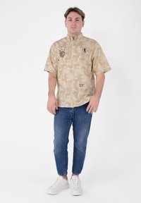 Camicia a maniche corte beige con motivo, grafica audace, bottoni e colletto, abbinata a jeans blu e sneakers bianche.