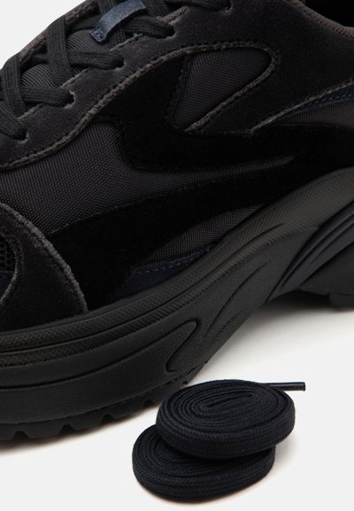 Gros plan d'une basket noire en tissu texturé avec des détails en daim, présentée avec une paire de lacets noirs enroulés sur une surface blanche.
