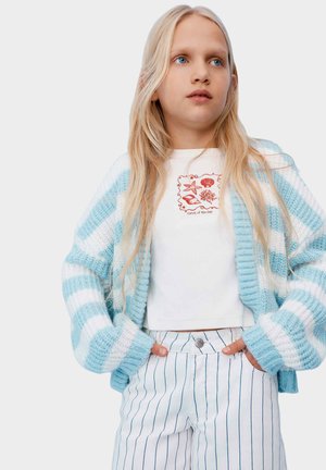 Blond meisje draagt een blauw-wit gestreepte gebreide cardigan, een wit crop top met rode zee-thema print en witte broek met blauwe verticale strepen.