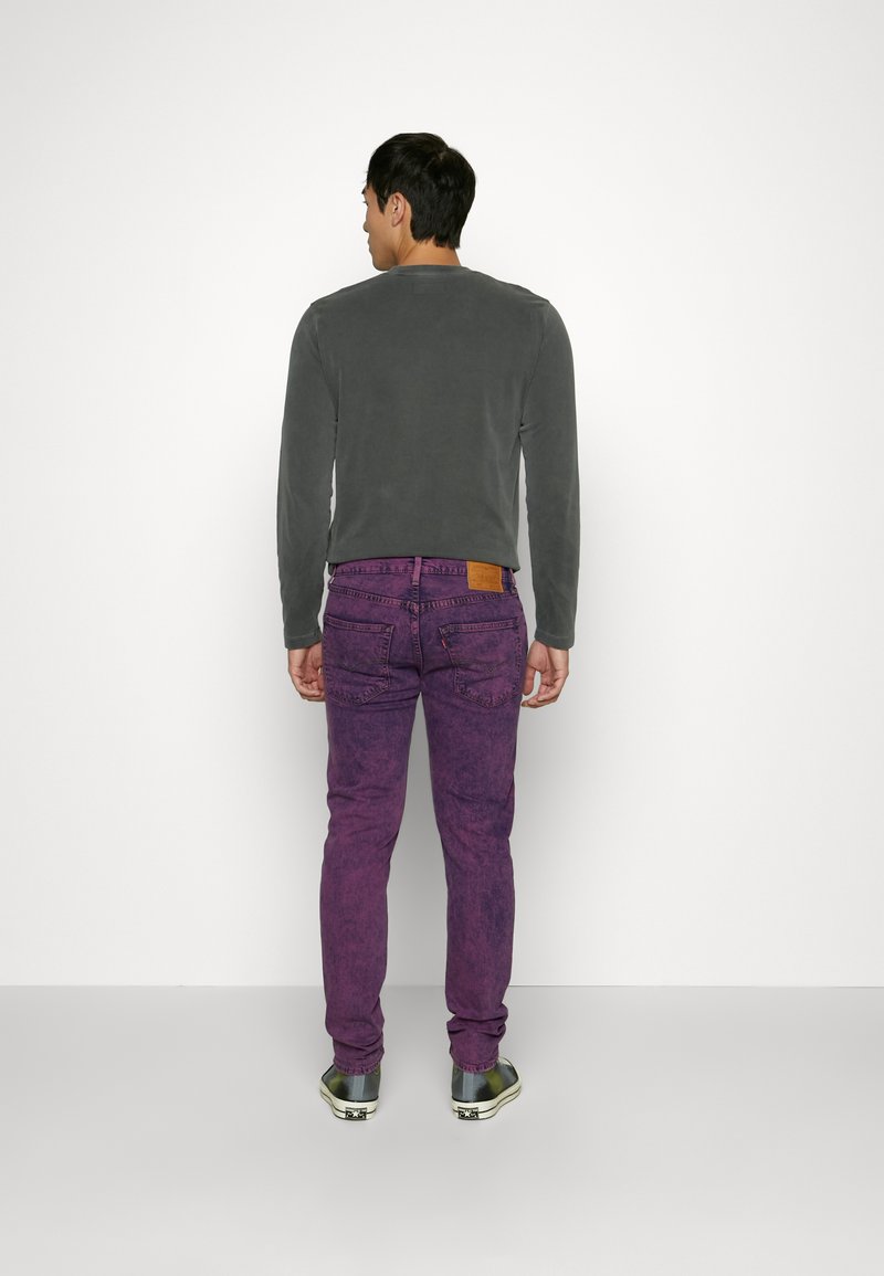 Camisa gris de manga larga, jeans morados con textura desgastada, parche de cuero marrón en la cintura y zapatillas grises. Vista trasera.