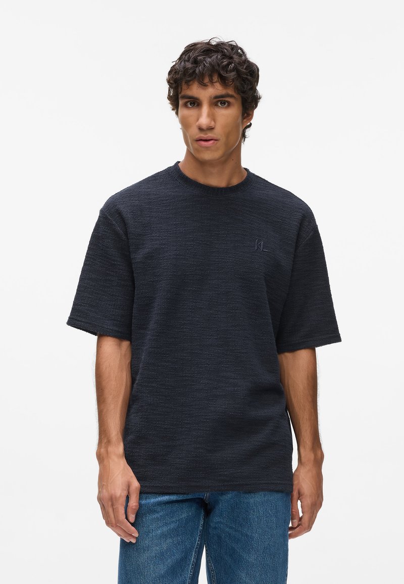 Dunkles, kurzärmliges, strukturiertes T-Shirt mit Rundhalsausschnitt und kleinem gestickten Logo. Kombiniert mit blauen Denim-Jeans, die eine lockere Passform bieten.