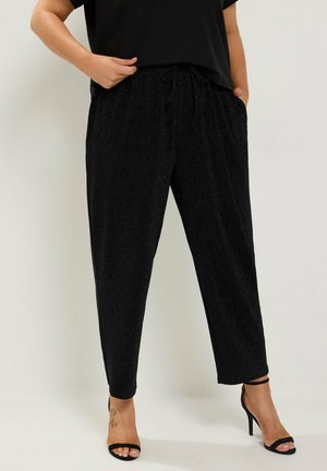 Donna che indossa pantaloni neri scintillanti a gamba affusolata con vita elastica e sandali neri con tacco alto, in piedi con le mani nelle tasche.