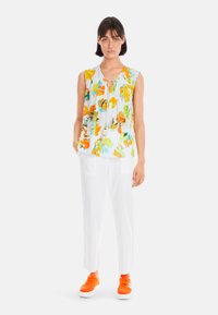 Weiße Bluse mit floralen Mustern in Orange und Grün, mit einem Schlüssel-Ausschnitt und Quasten-Akzenten, kombiniert mit weißen Hosen und orangenen Sneakers.