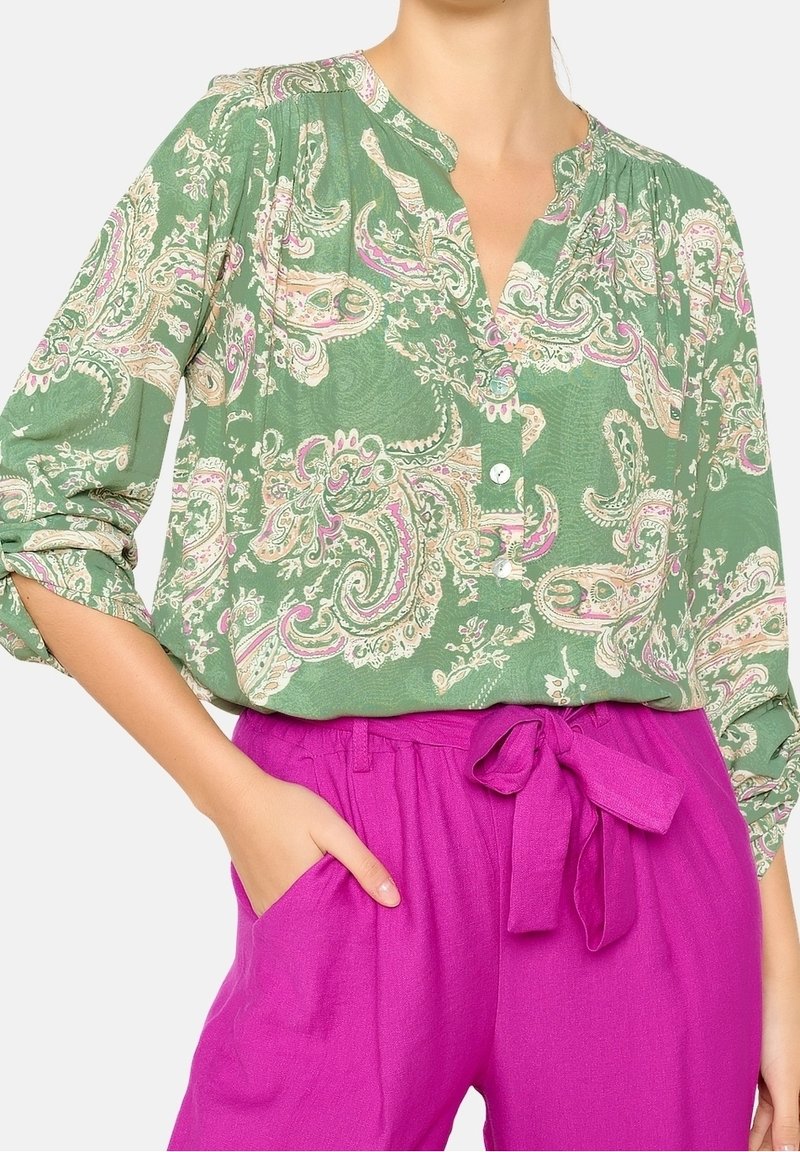 Blouse en paisley vert avec des manches retroussées, assortie à un pantalon fuchsia taille haute doté d'une ceinture en tissu nouée et de poches latérales.