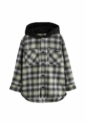 Persona vistiendo una camisa de cuadros beige y negra con botones, con dos bolsillos en el pecho, uno etiquetado como "SUSCRÍBETE", y una capucha negra debajo.