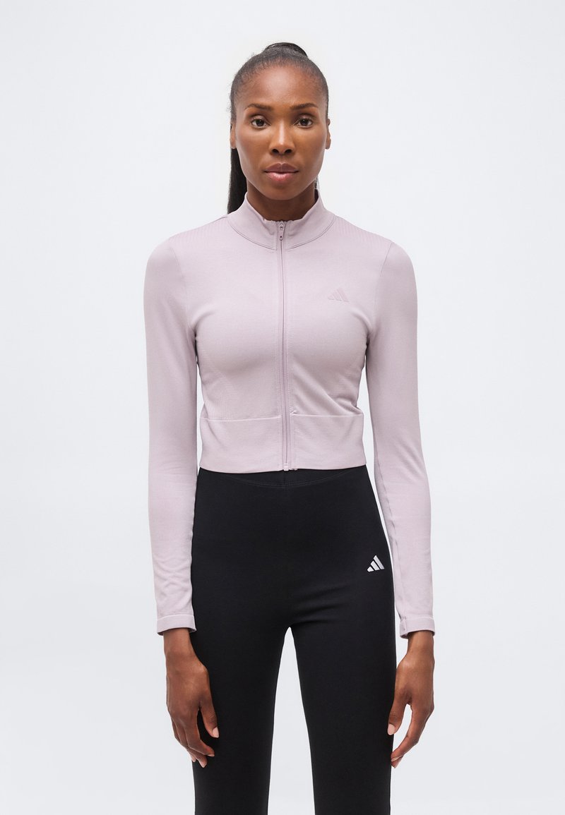 Femme debout, face à l'objectif, portant une veste zippée violet clair et des leggings Adidas noirs sur un fond blanc uni.
