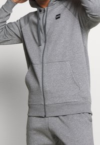 Sudadera gris con cremallera, hecha de una mezcla de algodón. Presenta un bolsillo frontal, capucha con cordón, puños acanalados y un logo en el pecho. Textura suave.