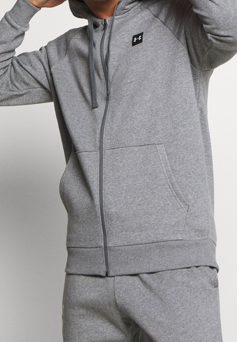 Sudadera gris con cremallera, hecha de una mezcla de algodón. Presenta un bolsillo frontal, capucha con cordón, puños acanalados y un logo en el pecho. Textura suave.