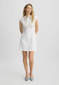 Abito bianco a camicia con corpetto aderente caratterizzato da decorative aperture ad occhio, maniche corte e una chiusura frontale con bottoni. Tessuto dalla consistenza liscia.