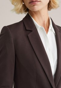 Hnedý blazer so štruktúrovaným dizajnom, so zárezovými klopami a hladkou textúrou, nosený cez bielu košeľu s golierom a gombíkmi.