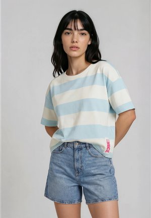 Vrouw met zwart haar, gekleed in een lichtblauw-wit gestreept shirt met korte mouwen en een spijkershort, staand met de handen achter de rug.